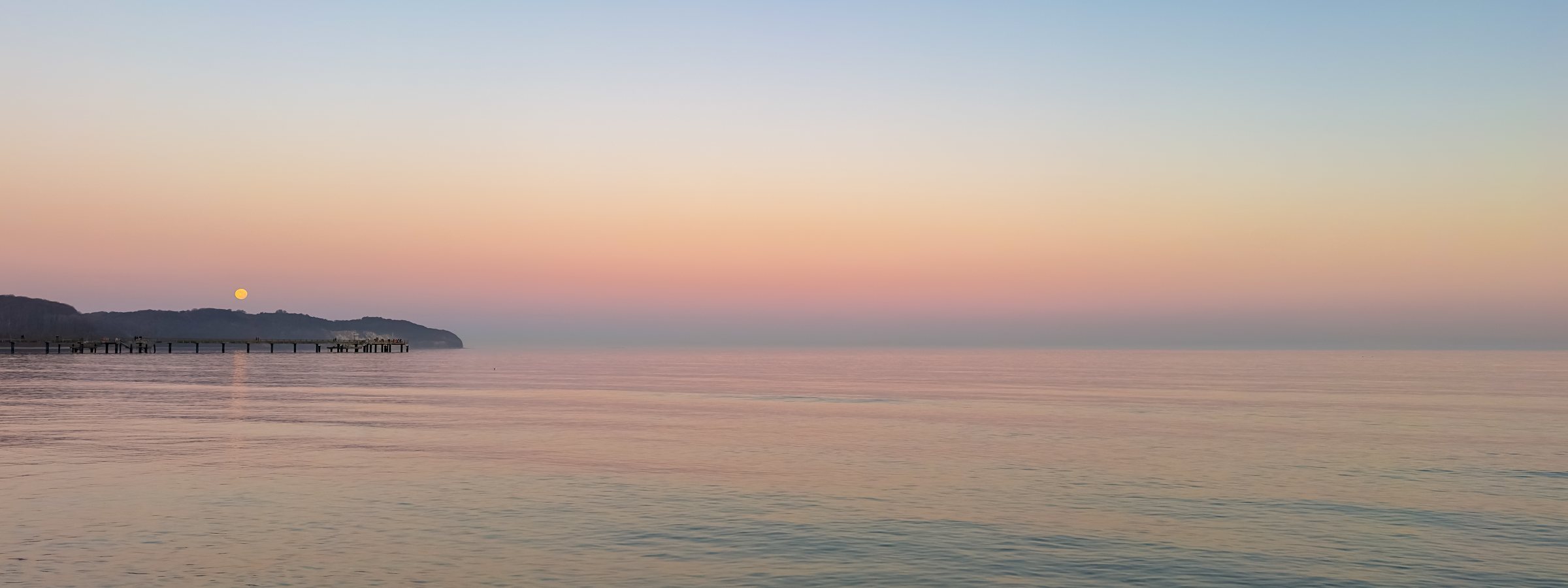 Calm sunset over the sea with a wooden pier and full moon. Peaceful twilight landscape.