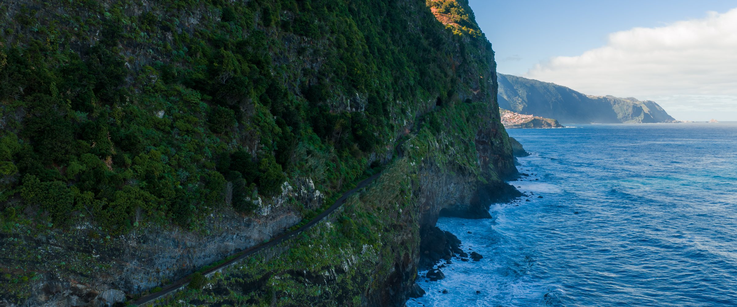 Late afternoon light warms basalt cliffs on Madeira, Portugal, as waves crash below a narrow road above the Atlantic, conveying isolation and wild coastal adven