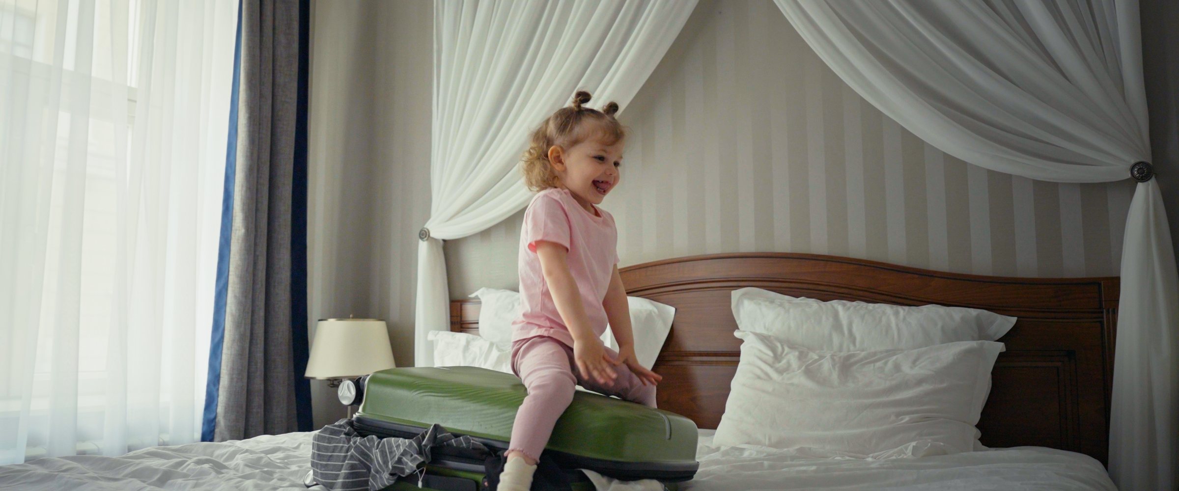 Excited little girl sitting on a half packed green suitcase on a white bed, smiling with anticipation as she prepares for a family trip, holiday or overnight ad