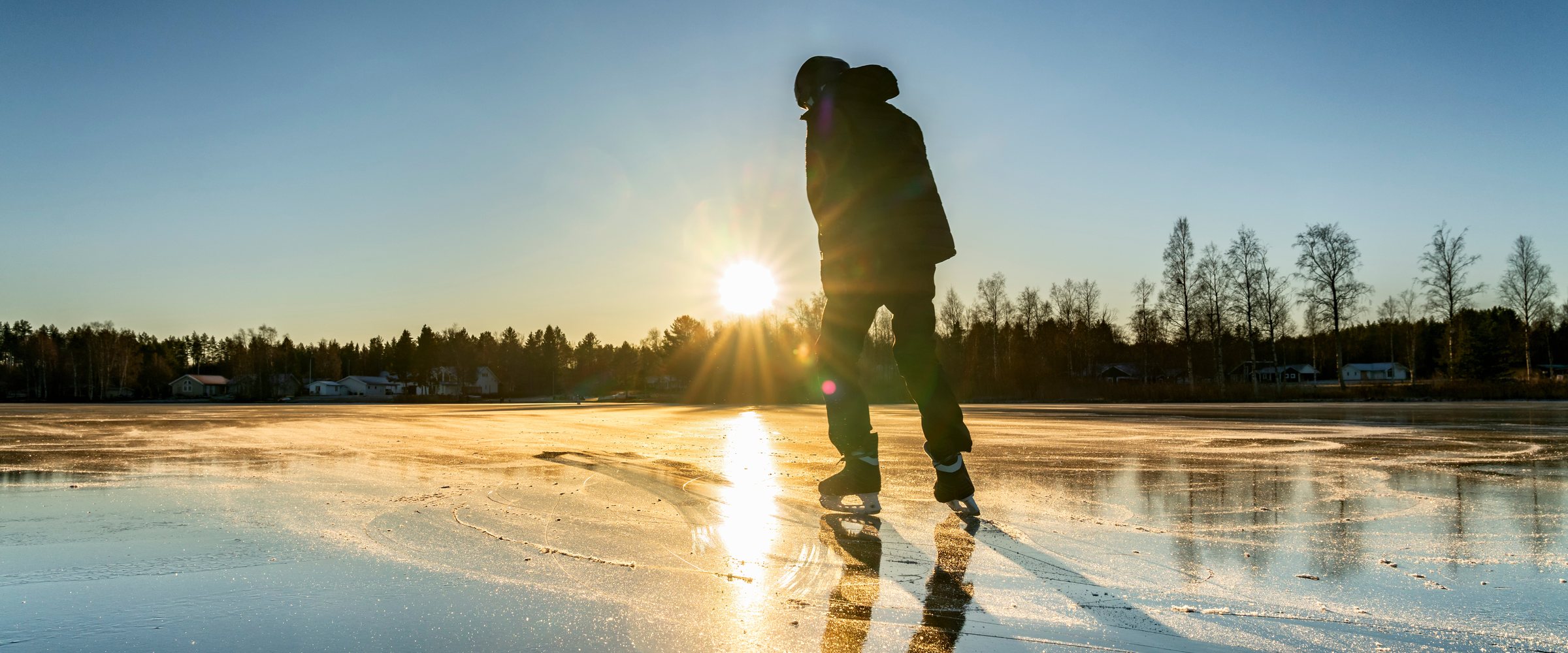 Boy silhouette against of the Sun: a teenager is ice skating on the crystal clear frozen lake - ice surface like big mirror. Low sun lights with warm light at v