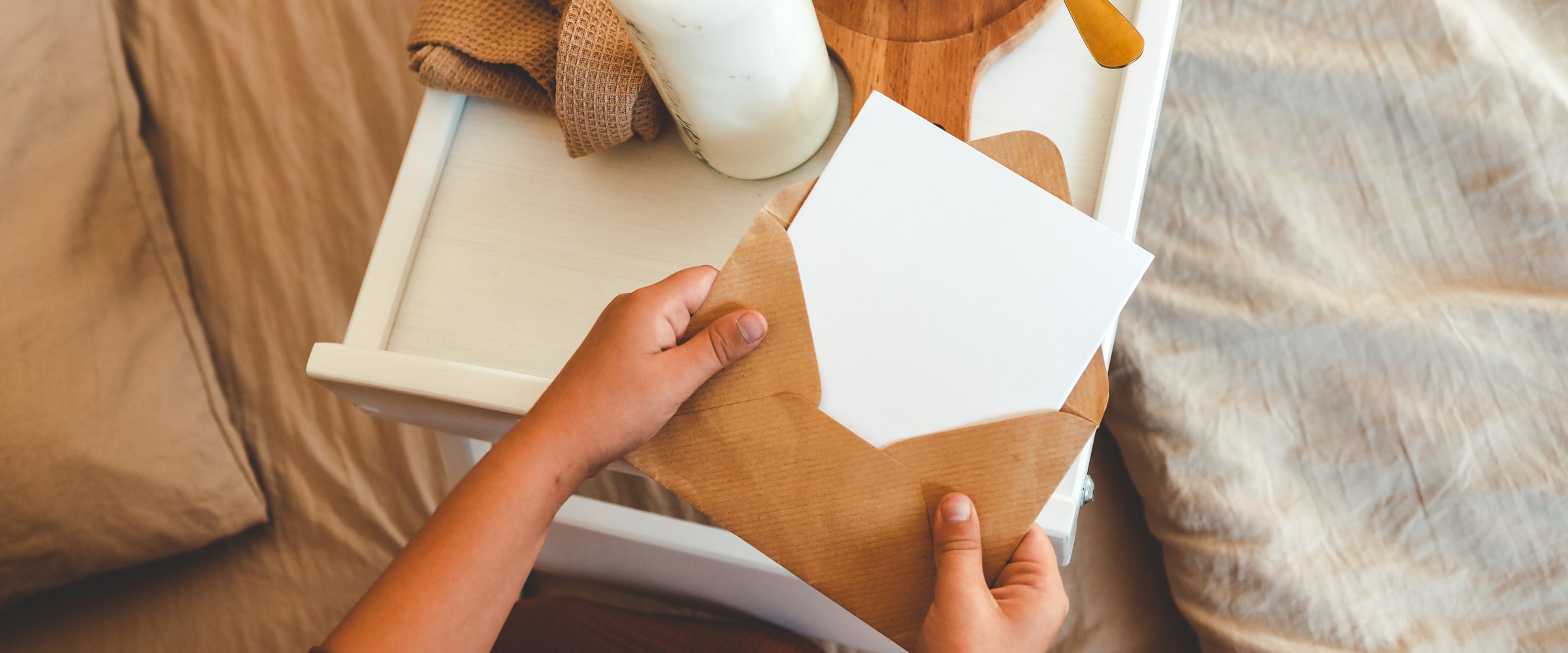 Child opening kraft envelope with card on a cozy bed, next to breakfast tray with granola and milk, and a wrapped gift box. Peaceful and warm morning celebratio